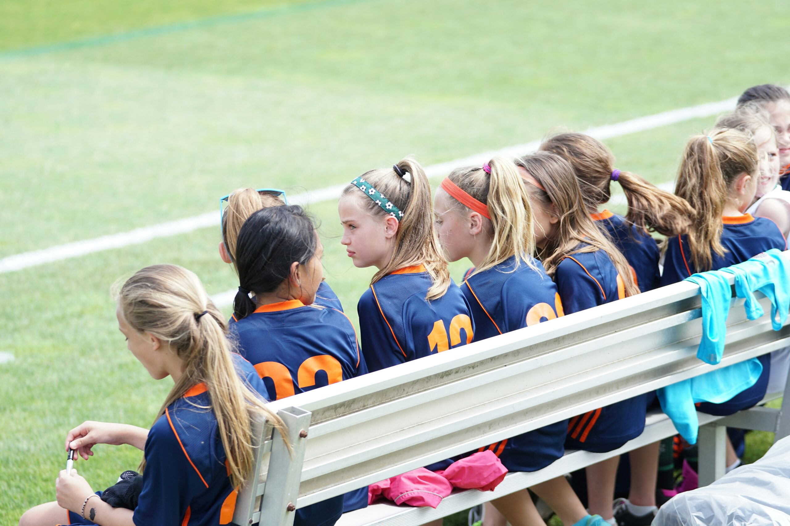 Young girls in blue and orange soccer uniforms sitting on a bench at a sports field