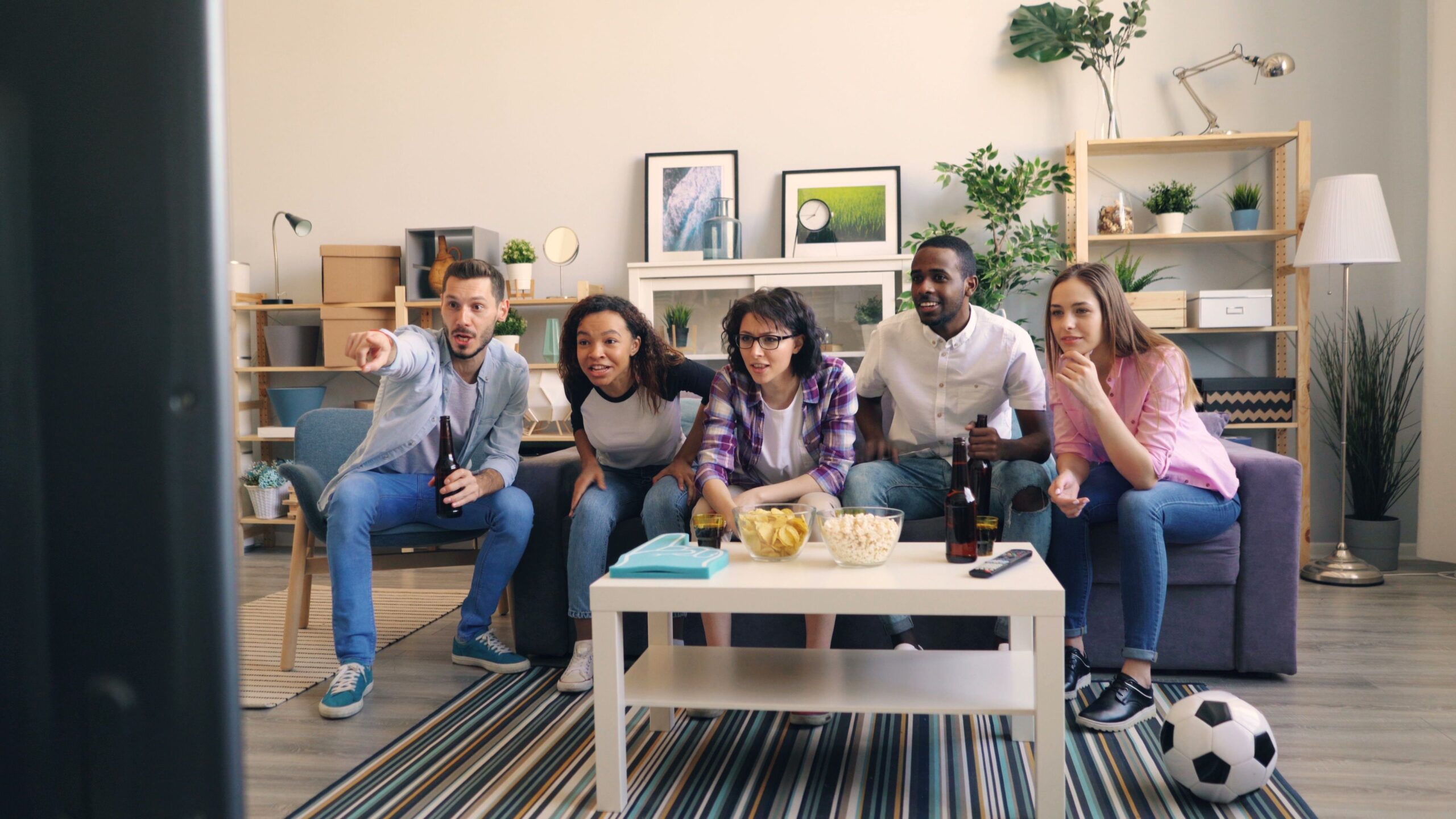 Five young adults watching TV intently, with snacks and drinks on a coffee table