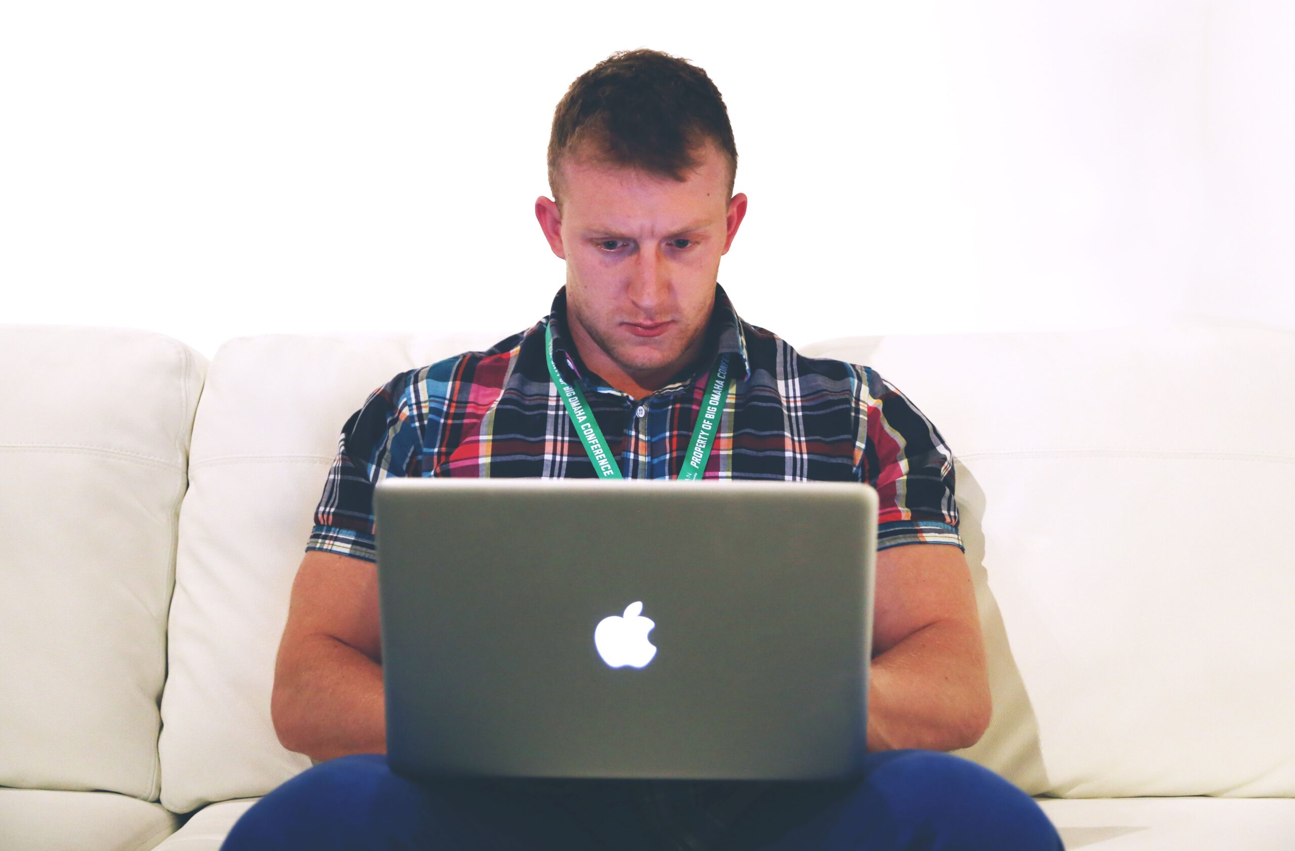Man in plaid shirt and blue pants sitting on a white couch using a laptop