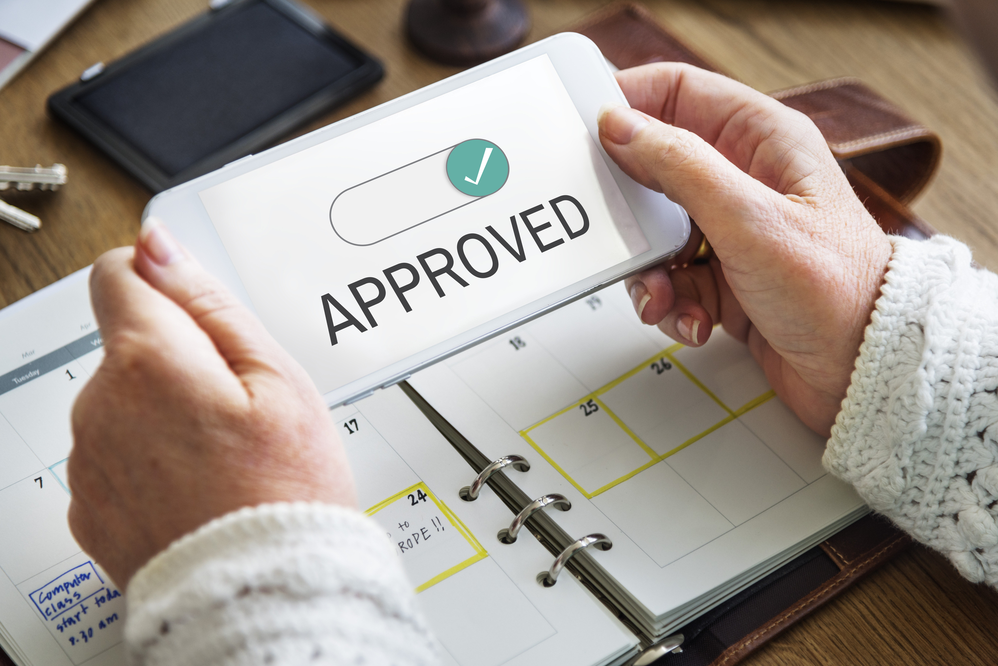 Hands holding a phone displaying "APPROVED" over an open calendar and wooden desk.